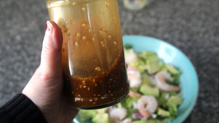 A jar filled with Sesame Ginger Dressing is held in a hand poised above a salad in a bowl on a granite countertop.