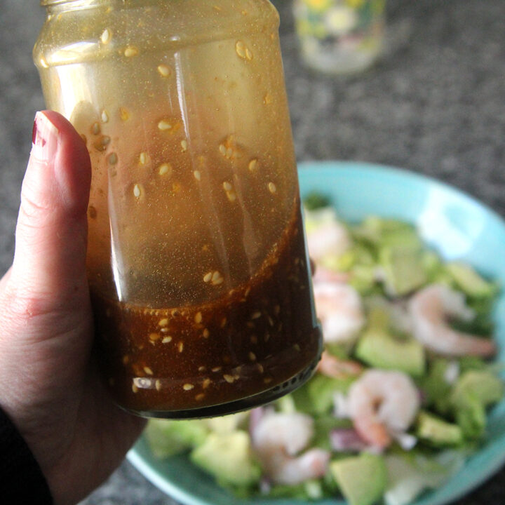 A jar filled with Sesame Ginger Dressing is held in a hand poised above a salad in a bowl on a granite countertop.