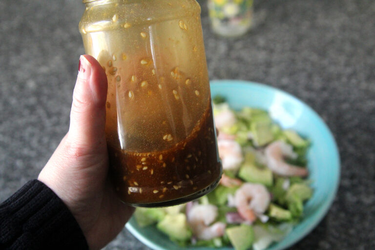 A jar filled with Sesame Ginger Dressing is held in a hand poised above a salad in a bowl on a granite countertop.
