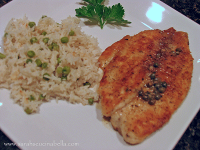 A white, square plate holds an oblong piece of golden brown fish with visible green capers on top. It's glistening. Next to it is a pile of creamy-colored rice topped with scallions. A leaf of parsley can be seen in the background.