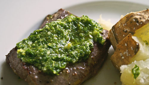 This photo shows a grilled steak on a white plate topped with green Lemon Parsley Sauce. There's also potatoes on the plate.