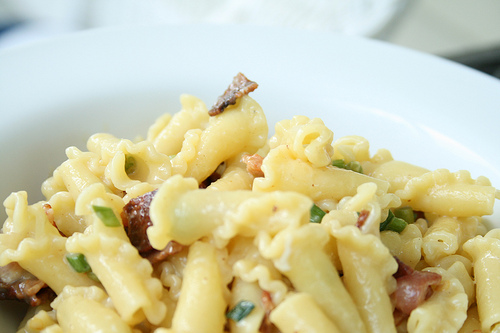 A white bowl featuring pasta with bacon, a creamy sauce and little bits of green garlic scapes.