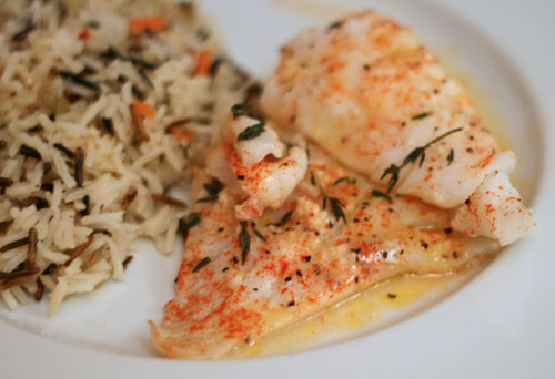 Cod, dusted with red paprika and thyme leaves, is on a white plate with rice pilaf piled nearby.