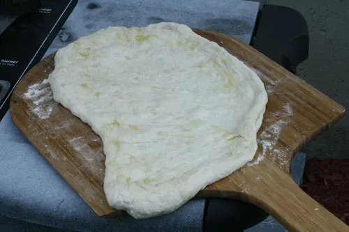 A pizza crust is spread out on a floured pizza peel with visible yellow oil on top.
