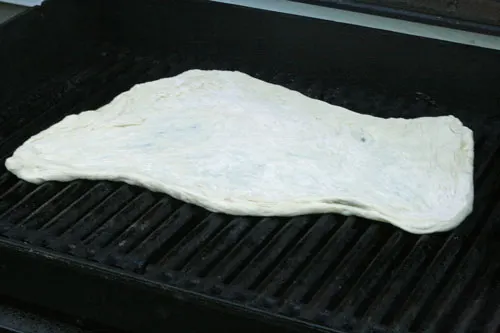 The pizza crust is spread in an irregular oblong shape on the black grates of a grill.