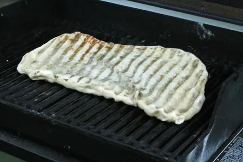 The pizza crust is spread in an irregular oblong shape on the black grates of a grill.