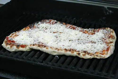 The pizza crust is spread in an irregular oblong shape on the black grates of a grill and spread with red sauce and white cheese.