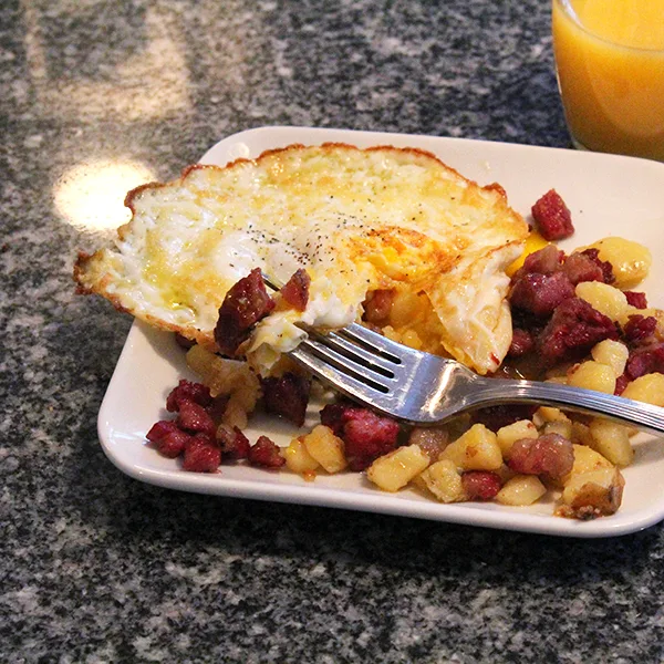 A square white plate of corned beef hash with a fried egg sits on a granite countertop with a forkful of hash resting on the plate.