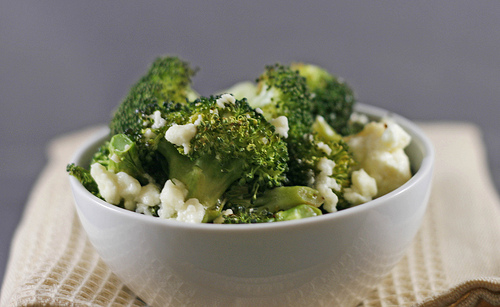A white bowl set on a tan towel holds roasted broccoli dotted with feta and garlic.