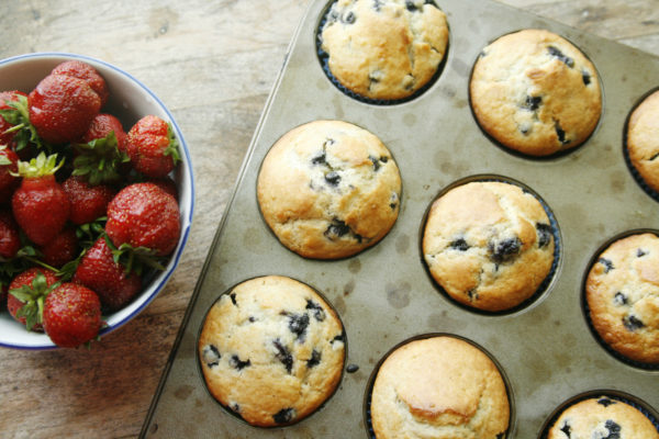 This shows a muffin pan of blueberry muffins with a bowl of strawberries to illustrate the Best Blueberry Muffins Recipe