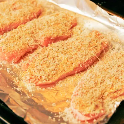 A foil-lined baking sheet holds four turkey cutlets covered with breadcrumbs, cheese and more.