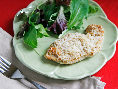 A Rosemary Romano Roasted Turkey Cutlet sits on a green plate with salad nearby. The table is covered in a red tablecloth and a tan napkin and fork sit near the plate.