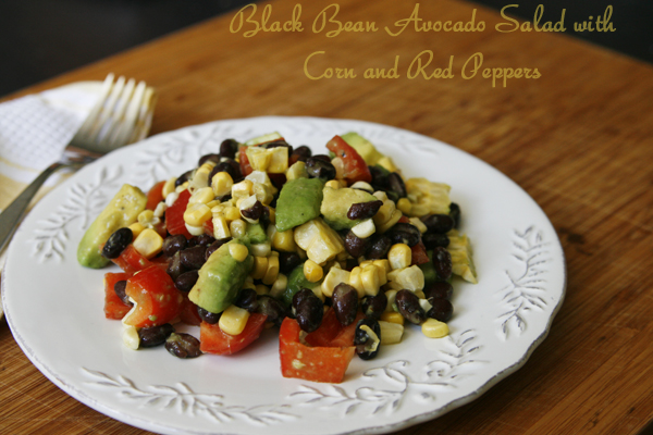 Black Bean Avocado Salad with Corn and Red Peppers is shown on a white plate on a wooden surface with a napkin and fork nearby.