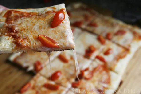 A slice of pizza is elevated from a rectangular pizza with cheese threading down.