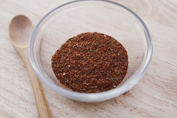 A glass bowl of red-toned spices on a wooden surface with a wooden spoon in the background illustrate this quick homemade taco seasoning recipe
