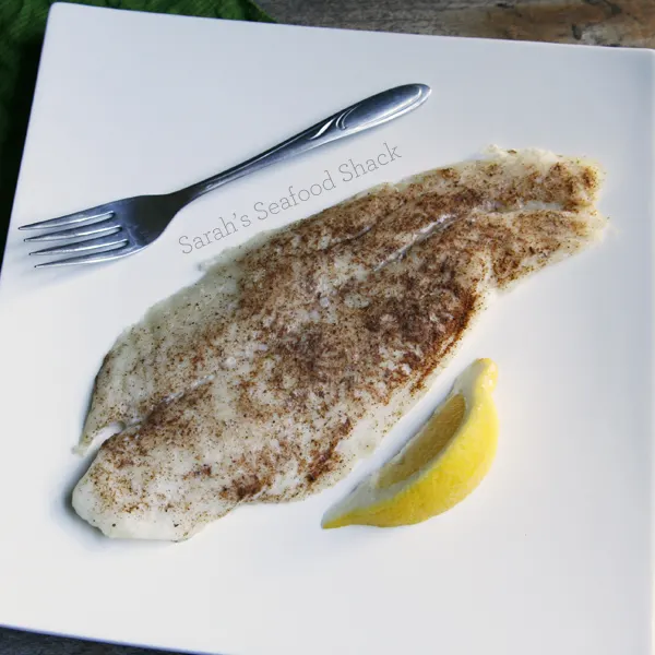 A white fish with orange and brown seasoning is shown on a white plate with a fork and a slice of lemon.