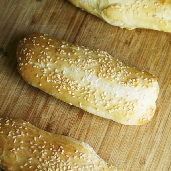 This photo shows homemade hoagie rolls on a wooden cutting board to illustrate this Sesame Hoagie Roll Recipe.