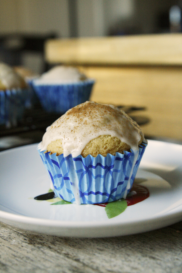 A light colored cupcake with a glaze dripping down over the blue cupcake paper is shown on a white plate. A sprinkle of cinnamon tops the cupcake. 