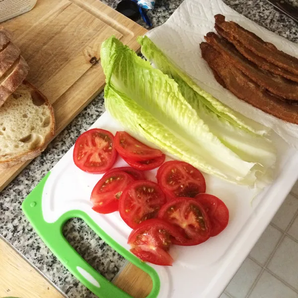 This shows the ingredients for a BLT on a white cutting board: tomatoes, lettuce and bacon. Sliced bread sits on a wooden cutting board nearby.