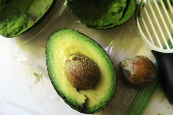 A green avocado is shown on a wooden cutting board with more avocado skins, an avocado pit and an avocado tool nearby.
