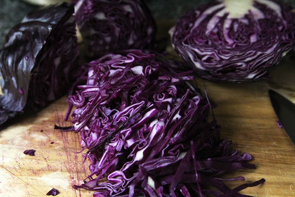 Thinly sliced red cabbage is shown on a wooden cutting board with a half-red cabbage and a quarter-red cabbage nearby. There's also the blade of a large knife visible.