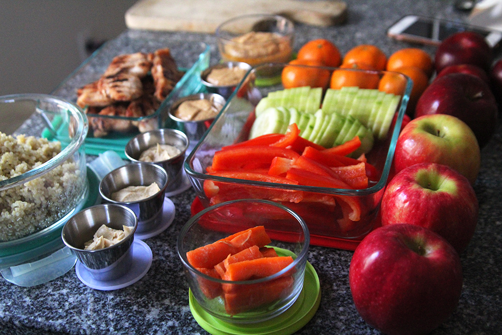 A granite countertop is shown covered with glass and metal containers containing hummus, vegetables, chicken, quinoa and sauces. There are also apples and clementines nearby.