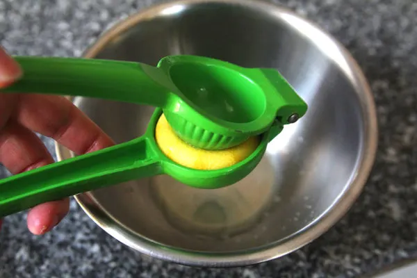 A lemon is being squeezed in a green citrus juicer over a silver mixing bowl.