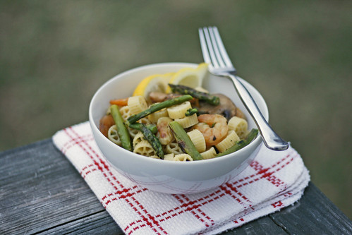 A white bowl on a white and red napkin sits on wood. Inside the bowl are green asparagus pieces, pink shrimp, yellow lemon wedges and cream colored wheel-shaped pasta.
