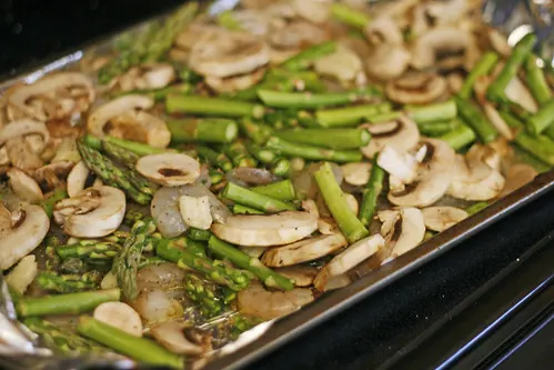 A baking sheet lined with silver aluminum foil is topped with green asparagus, tan mushrooms and white shrimp.