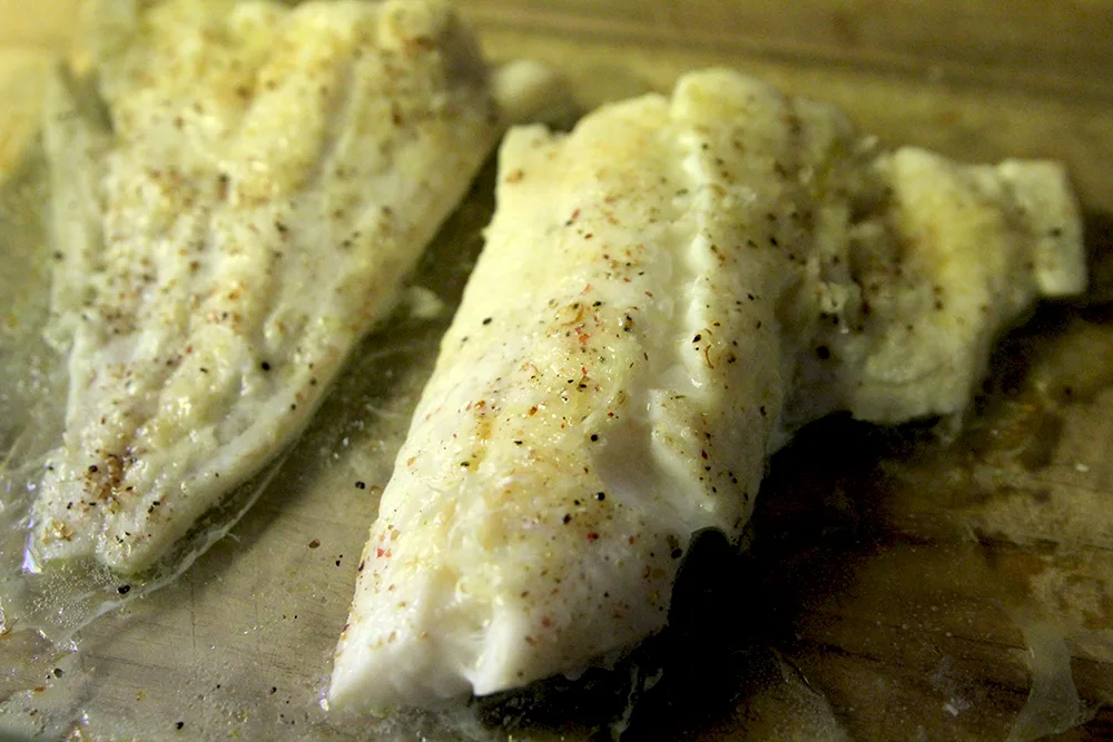 Fresh from the oven roasted haddock sits in a glass pan on a wooden surface. There is visible seasoning on the fish.