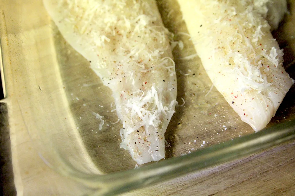 Two haddock fillets with visible seasoning and grated cheese sit in a glass pan on a wooden surface.