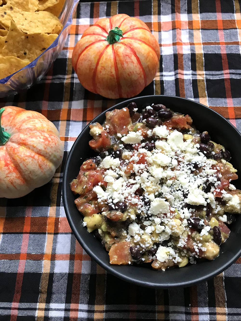 Black Bean, Tomato, Avocado and Cotija Cheese Dip in a black bowl on an orange, black and white plaid cloth. Two small pumpkins are nearby.