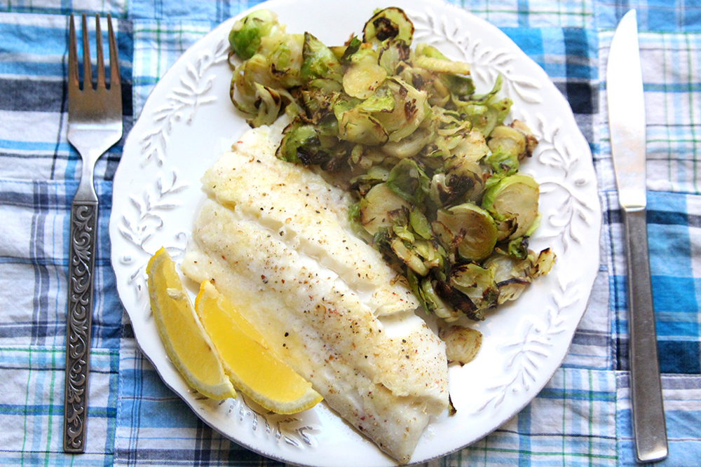 Parmesan Baked Haddock is shown on a white plate with a pile of sauteed Brussels sprouts and two lemon wedges. The plate sits on a blue plaid fabric-covered table with a fork and knife on either side.
