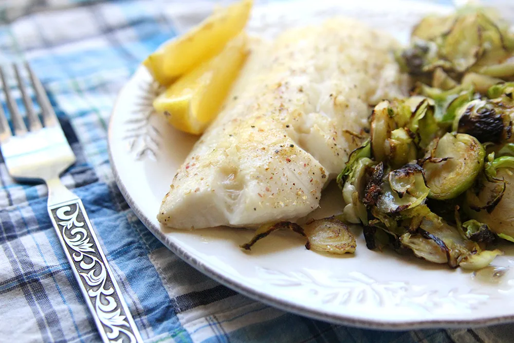 Parmesan Baked Haddock is shown on a white plate with two lemon wedges and a pile of cooked Brussels sprouts. The plate sits on a blue plaid fabric-covered table with a fork nearby.