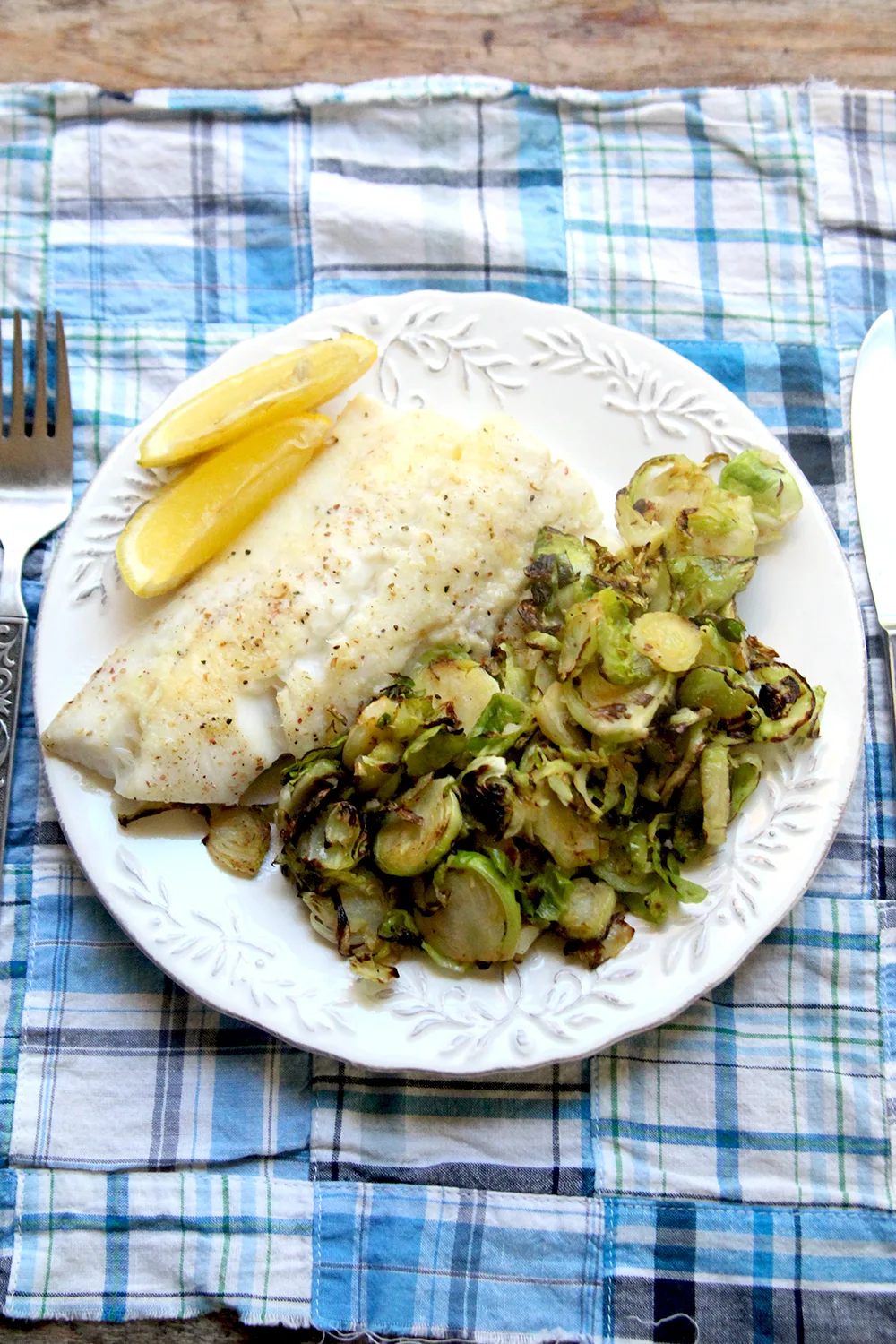 Parmesan Baked Haddock is shown from above on a white plate with a pile of sauteed Brussels sprouts and two lemon wedges. The plate sits on a blue plaid fabric-covered table with a fork and knife on either side.