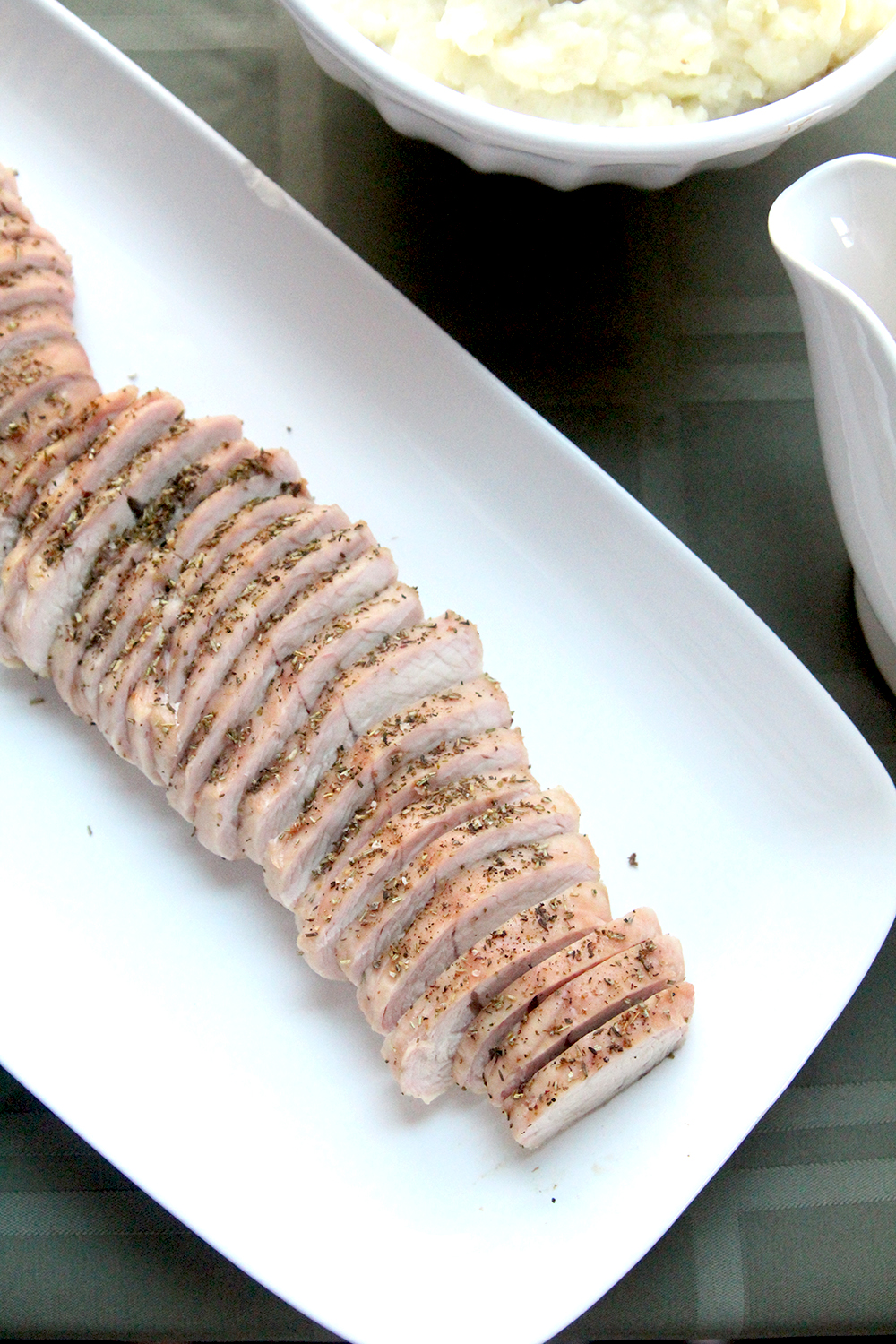 Roasted turkey tenderloin is shown from above, sliced, on a white platter with a white bowl of mashed potatoes and a gravy boat nearby. There's a green tablecloth on the table. 