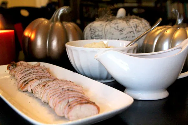 Roasted turkey tenderloin is shown, sliced, on a white platter with a white bowl and a gravy boat nearby. There's a green tablecloth on the table and decorative pumpkins and candles in the background.