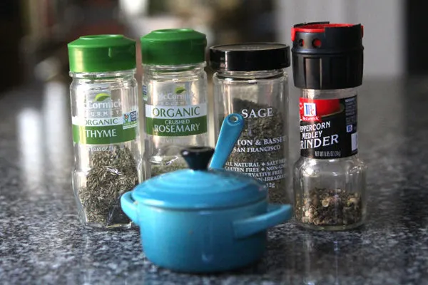 Four containers of herbs and spices are shown with a blue container on a countertop.