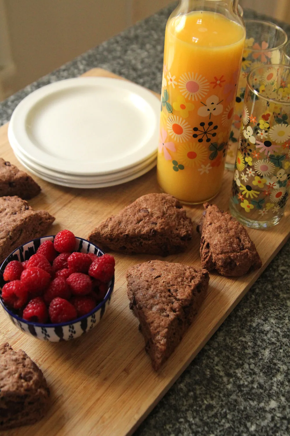 double chocolate scones recipe shown with a glass pitcher of orange juice, a bowl of raspberries and a stock of white plates on a wooden cutting board.