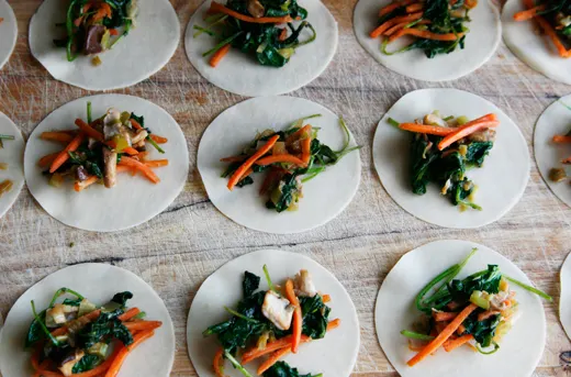 Dumpling wrappers topped with green and orange veggies sit on a wood cutting board.