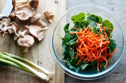 A bowl of kale and carrots sit near a cutting board of shiitake mushrooms and scallions.