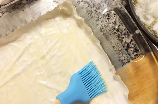 A blue brush spreads butter on phyllo dough in a glass dish.
