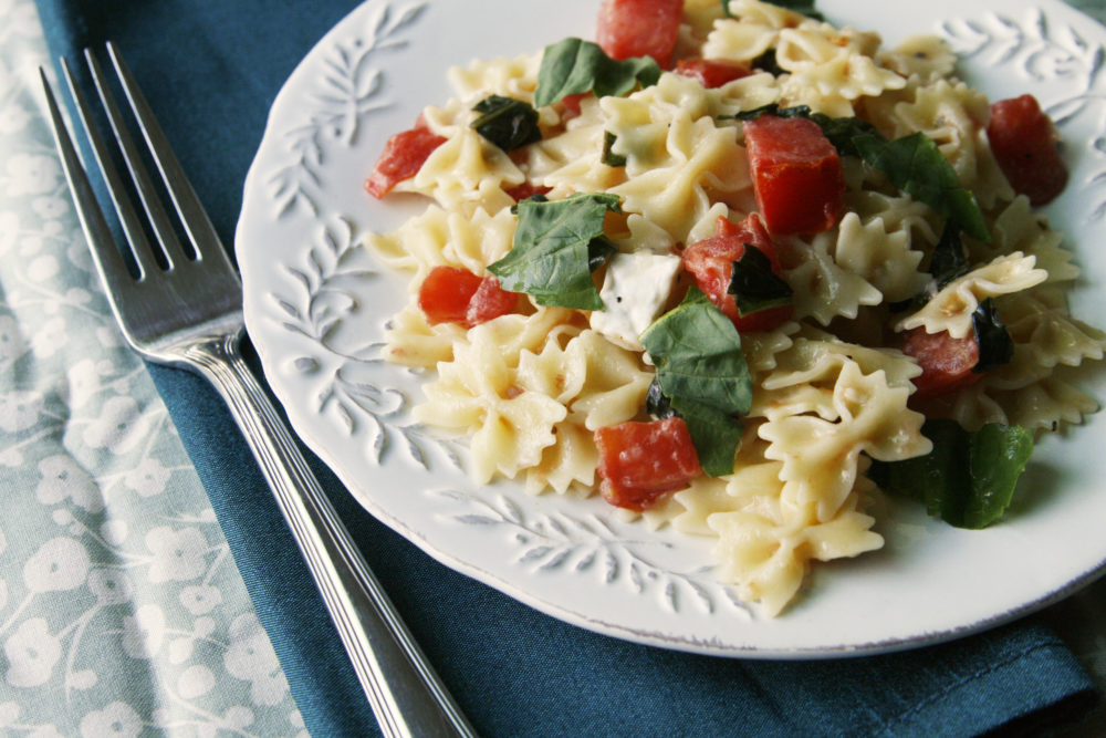 A white plate with bowtie pasta featuring tomatoes, basil and brie sits on a table with a fork.