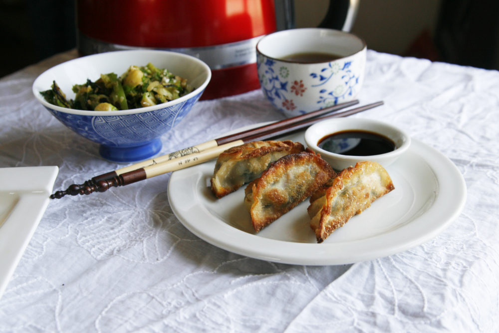A white plate with Kale and Mushroom Gyoza and a small bowl of soy sauce sit with chopsticks. A tea cup with tea and a bowl with green vegetables sits nearby.