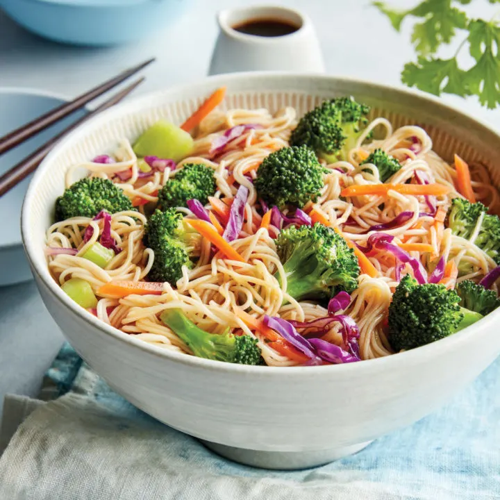 A white bowl has noodles with broccoli, red cabbage and other vegetables in it.