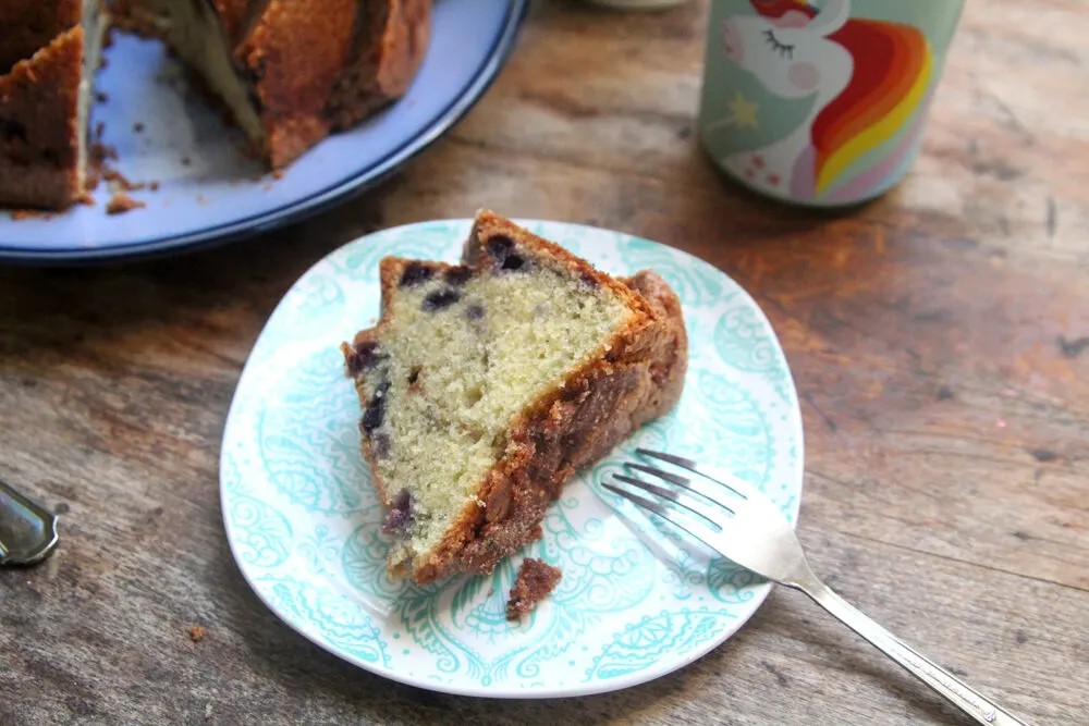 A slice of wild blueberry pound cake is on a plate with a fork. The rest of the cake and a unicorn measuring cup sit nearby.