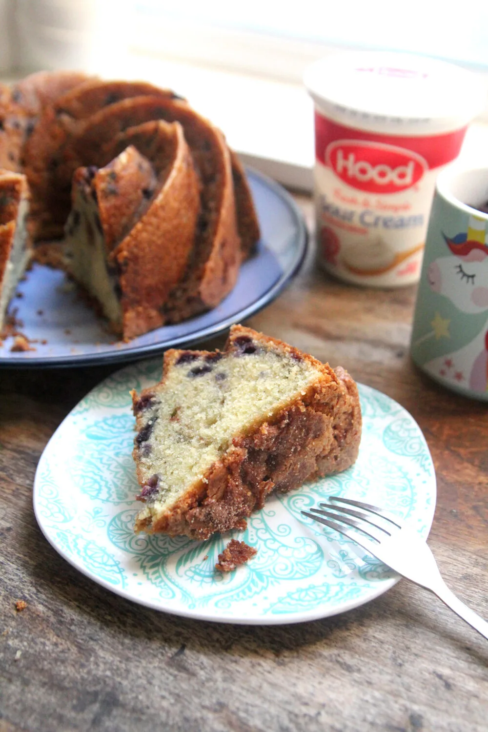 A slice of wild blueberry poundcake is on a small plate. The rest of the cake is on a big plate. Hood Sour Cream and a unicorn measuring cup sit nearby.