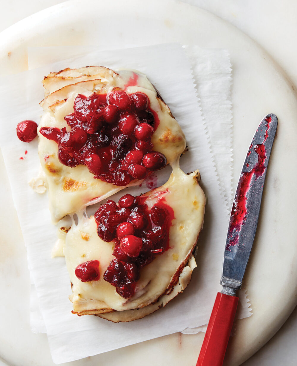 An Open-Face Turkey Cranberry Sandwiches with Pepper Jack Cheese sits on white parchment paper with a knife nearby. It's cut in half.