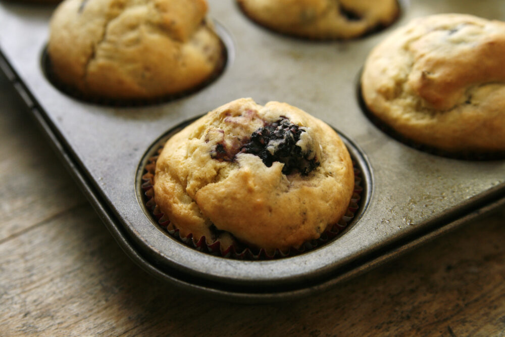 A muffin pan, showing signs of use, is shown on a wooden table. Inside the pan are Blackberry Ginger Muffins, hot from the oven.