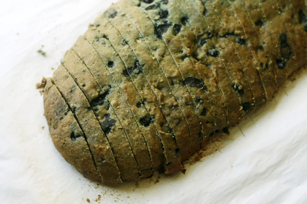 A browned loaf with dots of blueberries is shown sliced on parchment paper.